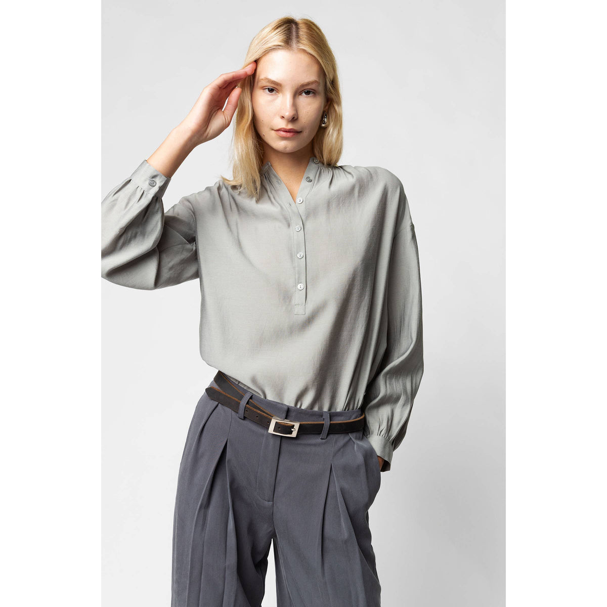 A woman wears an elegant, grey Naked Zebra - Button-down Blouse With Gathered Neckline: Black / L tucked into grey pleated trousers with a black belt.