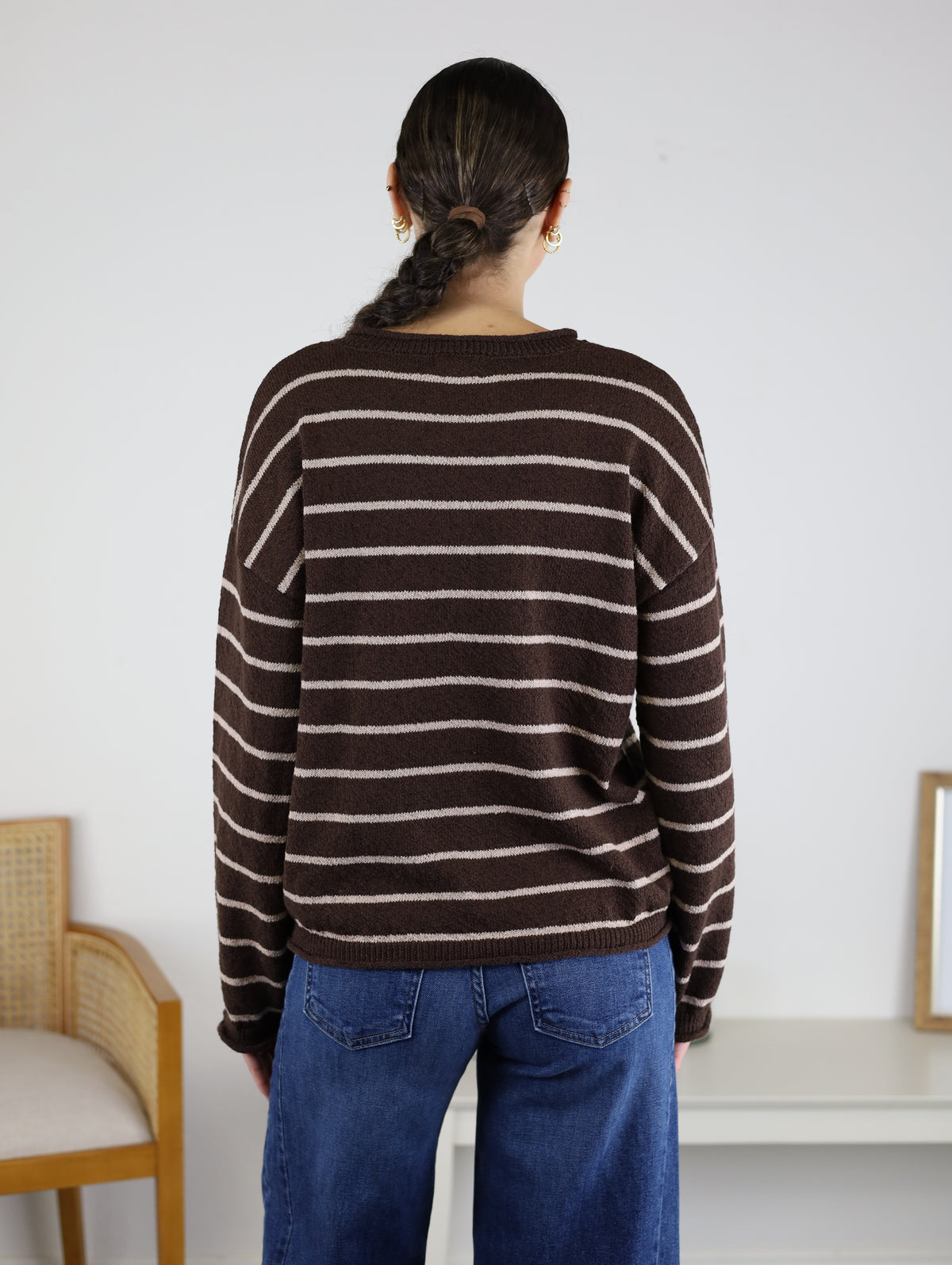 A person with dark braided hair, wearing the Odette Stripe Sweater—a relaxed pullover cotton knit in brown and white stripes—paired with blue jeans, stands facing away in a simple room furnished with a chair and a white table.