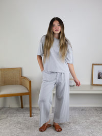 A woman stands indoors in the Alanna Striped Knit Top and Pants Set—light gray and white with wide-leg pants and a short-sleeve top—barefoot beside a wooden chair and low white table with framed pictures.