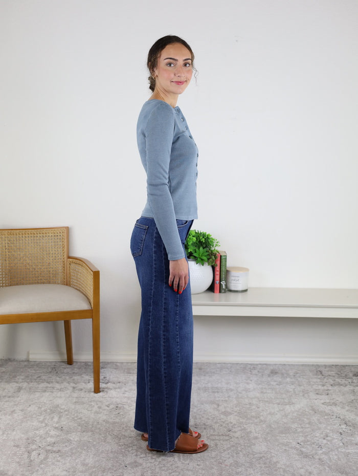 Wearing the Maisey Raw Hem Side Panel Wide Leg dark denim jeans and a blue long-sleeve top, a woman stands sideways in a room decorated with a chair, books, and a plant on a white shelf.