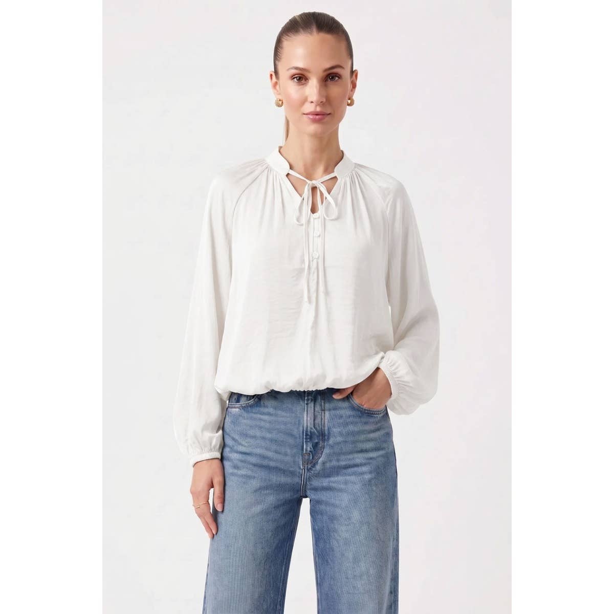 A woman wears a Naked Zebra - Billowy Tie Neck Long Sleeve Blouse: Chocolate / L, featuring a long-sleeved, off-white billowy design with a tie neck and blue jeans.