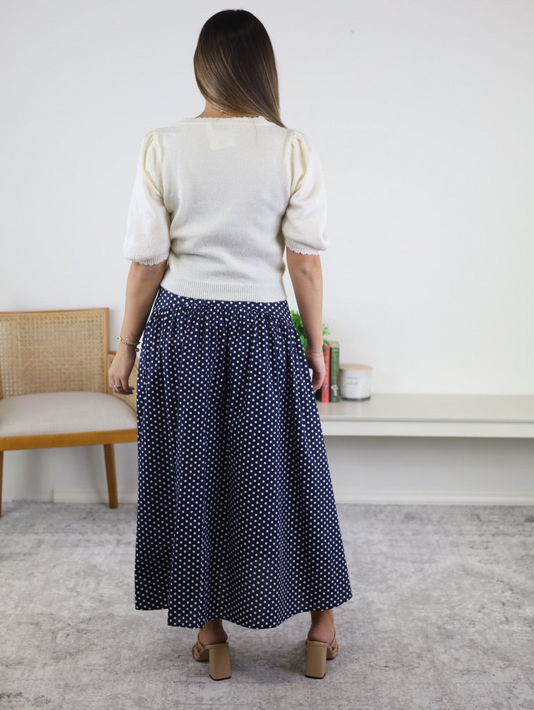 A woman with long hair stands indoors facing away, wearing the Monica Short Sleeve Cardigan in white, paired with a navy blue polka dot maxi skirt and sandals. A chair and decorative items are visible in the background.