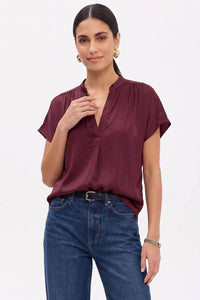 A woman wearing a dark red, short-sleeved Naked Zebra - Drop Shoulder Regular Blouse: Chocolate / L with a V-neckline and dark blue jeans.