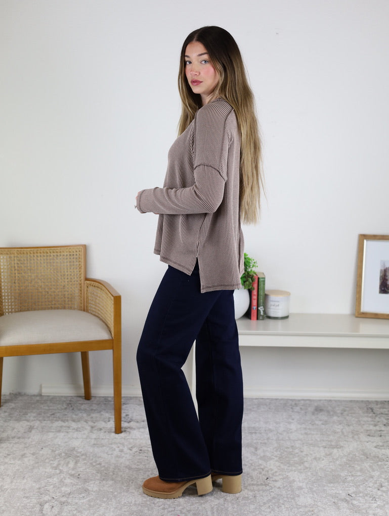 A woman with long hair stands indoors in a loose brown Finley Sweater with ribbed texture, dark wide-leg pants, and tan platform shoes. Behind her: chair, books, plant, and framed art.