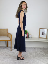 A woman wearing an Abigail Asymmetrical Shoulder Midi Dress in navy blue with a gathered waist detail, paired with black heeled sandals.
