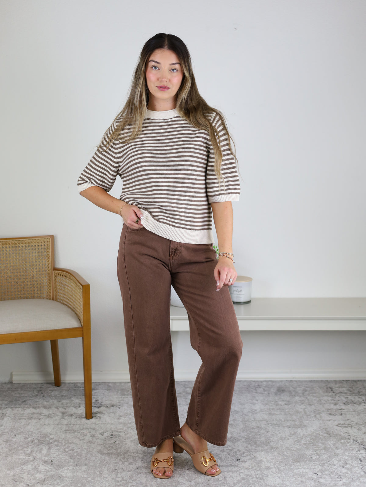 A woman stands indoors wearing the Scout Striped Sweater, a beige and brown striped top crafted from soft cotton knit. She pairs it with brown wide-leg pants and tan sandals, posing with one hand on her waist beside a wooden chair and a white bench against a plain wall.