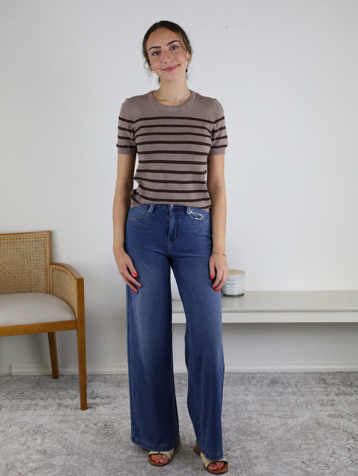 A woman stands indoors on a gray rug, wearing wide leg Rachelle Low Rise Denim jeans and a brown striped short-sleeve top. There is a wooden chair on the left and a white bench with a candle on the right against a plain white wall.