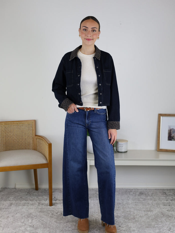 A woman stands indoors wearing the Autumn Leopard Collar Denim Shacket layered over a white top, paired with wide-leg blue jeans and brown shoes. She is positioned next to a chair and a shelf displaying a framed photo, candle, and other decorative items, against a plain background wall.