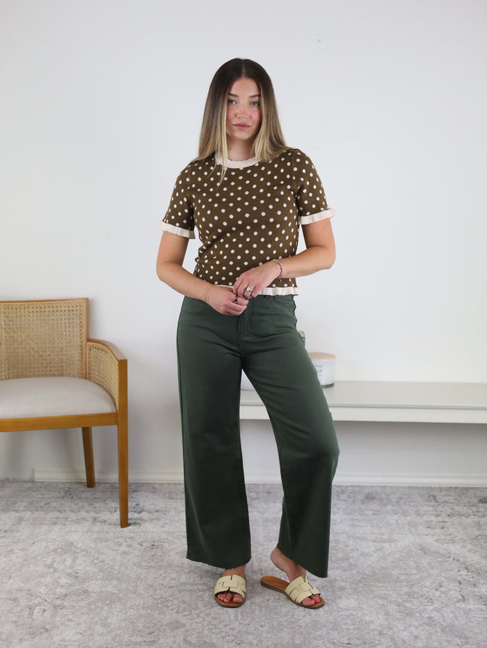 A woman stands indoors wearing the Lainey Polka Dot Sweater, paired with dark green wide-leg pants and cream sandals. She has straight hair and is positioned near a wooden chair and a white bench against a plain wall.