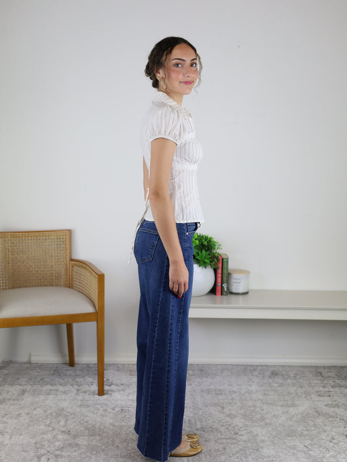 A woman stands sideways in a room, wearing the Cody Striped Blouse paired with wide-leg blue jeans. In the background, there’s a wooden chair, a white bench with books, a plant, and a candle.