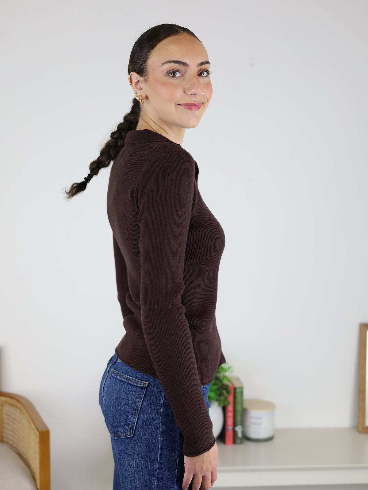 Wearing the Alexa Snap Button Ribbed Sweater, a person with dark braided hair and blue jeans stands sideways, smiling in a room decorated with a plant, books, and a candle on a white shelf.