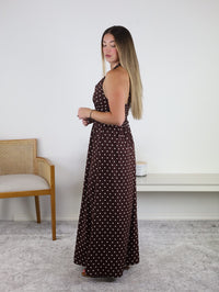 A woman stands sideways indoors near a chair and small shelf against a white wall, wearing the Marley Polka Dot Halter Maxi Dress in brown with white polka dots. Her long, straight hair complements this sleeveless resort-ready dress—an ideal addition to any vacation wardrobe.