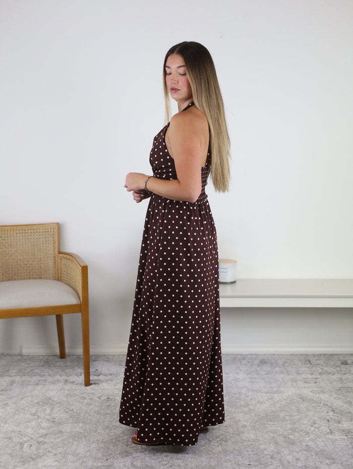 A woman stands sideways indoors near a chair and small shelf against a white wall, wearing the Marley Polka Dot Halter Maxi Dress in brown with white polka dots. Her long, straight hair complements this sleeveless resort-ready dress—an ideal addition to any vacation wardrobe.