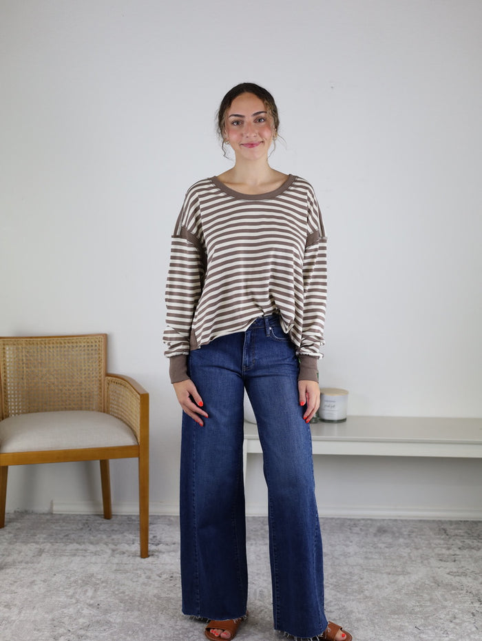 Woman standing indoors, wearing the Kiana Stripe Open Back Long Sleeve— a relaxed fit top with beige and white stripes—paired with wide-leg blue jeans and red sandals. A beige chair is to her left, and a white shelf with a candle is behind her.