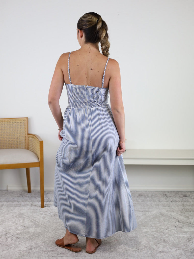 A woman with a braid stands indoors with her back to the camera, wearing the Molly Striped Maxi Dress—featuring blue and white stripes and adjustable straps—paired with sandals. A wooden chair and a white bench are visible in the background.