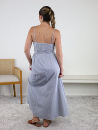 A woman with a braid stands indoors with her back to the camera, wearing the Molly Striped Maxi Dress—featuring blue and white stripes and adjustable straps—paired with sandals. A wooden chair and a white bench are visible in the background.
