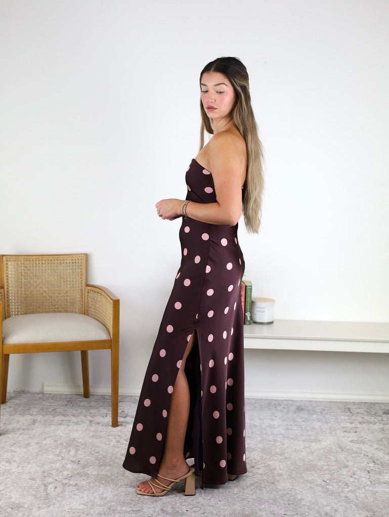 A woman stands sideways in the Polka Dot Satin Strapless Maxi Dress—a brown mauve maxi dress with pink polka dots and a side slit—paired with heeled sandals. She is in a minimalist room featuring a chair, a bench, and a few small items on the bench.