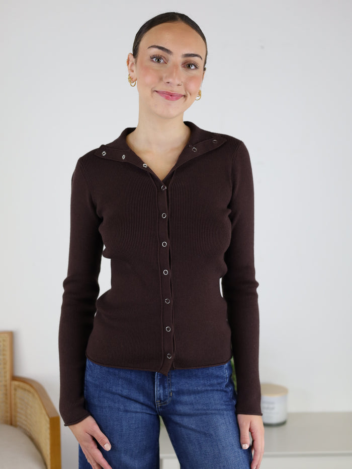 A woman with dark hair in a sleek bun wears the Alexa Snap Button Ribbed Sweater in brown along with blue jeans, standing indoors and smiling softly at the camera.