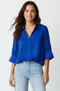 A woman wears a wine Naked Zebra - Half Sleeve Hi-lo Bubble Hem Blouse, featuring a collared V-neckline, half-length sleeves with pleated detailing, and a cobalt blue bubble hem.