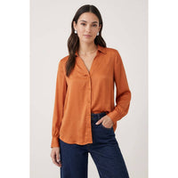 A woman wears a Naked Zebra - Neck Shirt With Open Seam Sleeve: Wine / L, a rust-orange, long-sleeved, collared button-down shirt with open seam sleeves, paired with dark blue jeans.