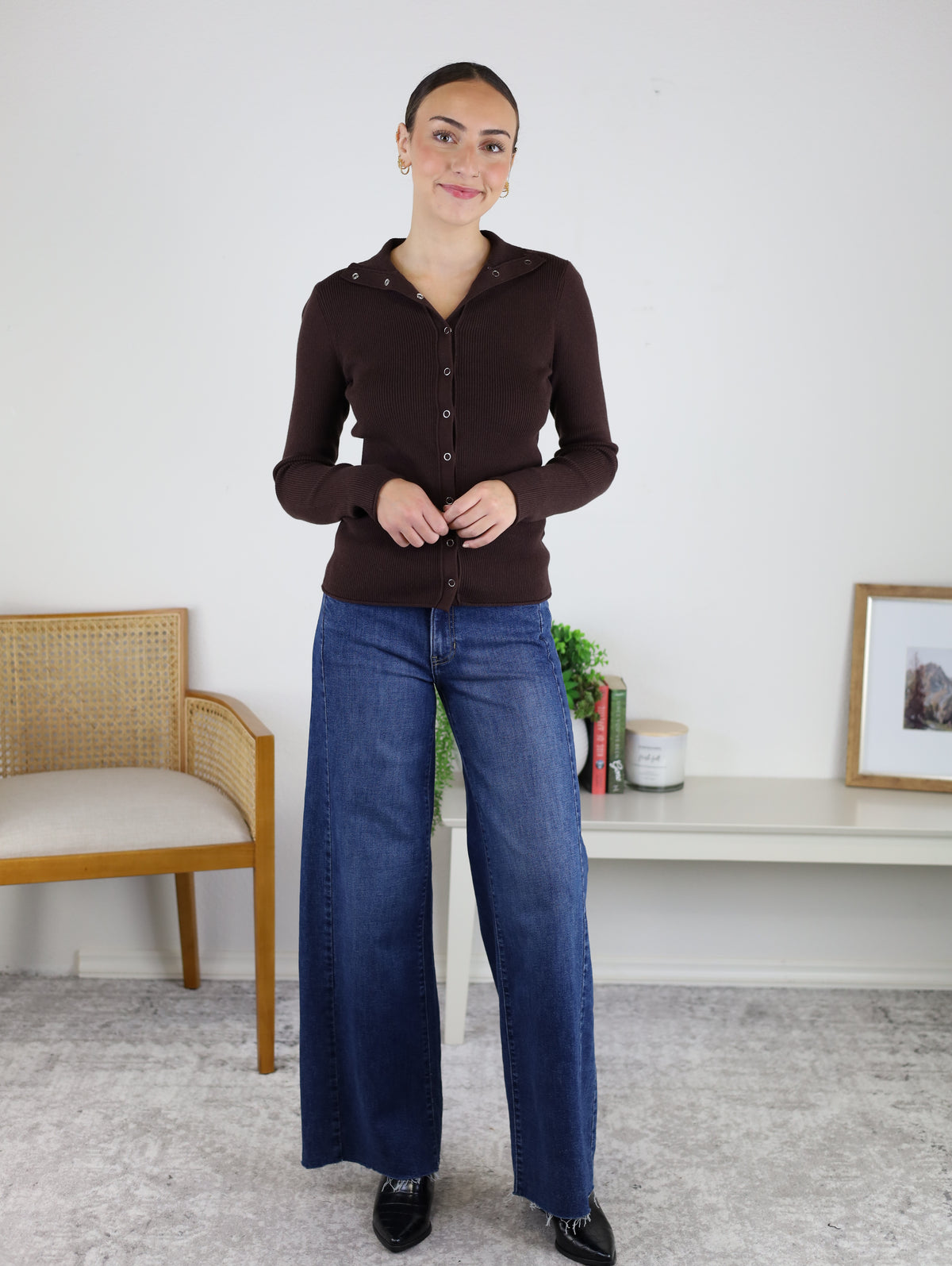 A woman stands indoors, smiling with her hands clasped, wearing the Alexa Snap Button Ribbed Sweater in dark brown paired with wide-leg blue jeans. Behind her are a wooden chair, a low white shelf adorned with decor items, and a framed photo.