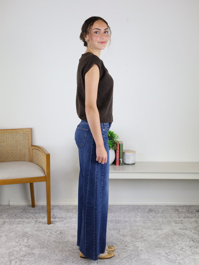 Woman standing sideways indoors, wearing a sleeveless brown button-down top, blue wide-leg jeans, and tan shoes. There is a wooden chair and a shelf with books, a plant, and a candle in the background. The sleeveless button-down is perfect for layering under the Camden Lightweight Cardigan or wearing on its own.