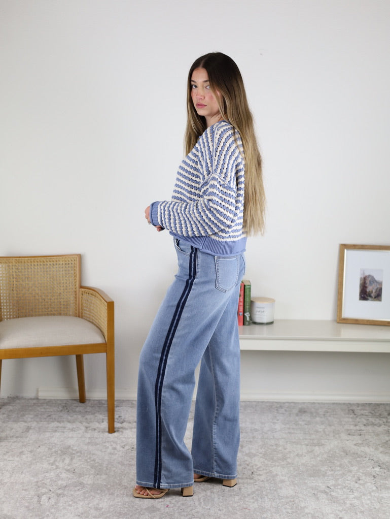 A woman with long hair wears the Bellamy Textured Striped Crochet Cardigan and wide-leg jeans with a dark side stripe, standing indoors on a light carpet near a chair and bookshelf.