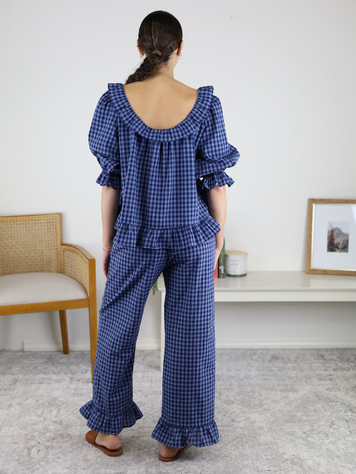 A woman stands with her back to the camera, wearing the Barron Cotton Gingham Frill Set in blue, which includes a frill top and matching pants. She is indoors, positioned near a wooden chair and a shelf adorned with decor items and a framed picture.