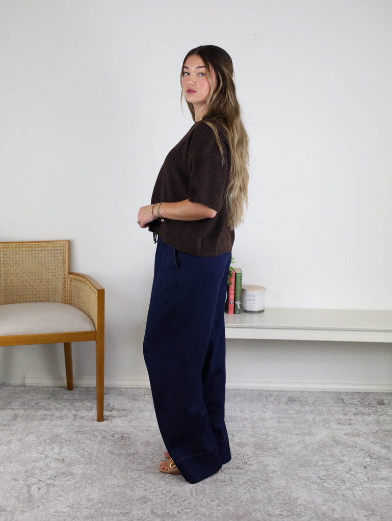A woman with long hair stands sideways in a room, wearing the West Raw Edge Sweater in brown paired with wide navy pants. Behind her are a wooden chair and a white shelf displaying books and a candle, set against a plain white wall.