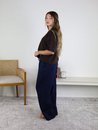 A woman with long hair stands sideways in a room, wearing the West Raw Edge Sweater in brown paired with wide navy pants. Behind her are a wooden chair and a white shelf displaying books and a candle, set against a plain white wall.