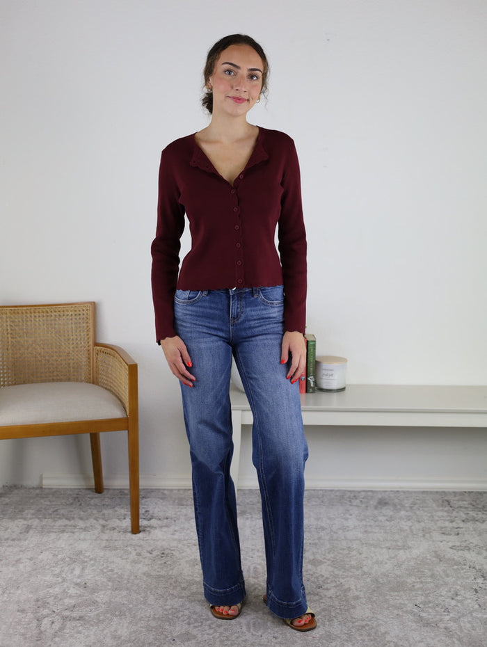 A woman stands indoors, wearing the Juniper Scalloped Cardigan in burgundy, paired with blue jeans and sandals. She is next to a chair and a white shelf with candles against a plain white wall.