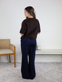 A woman with long hair stands facing away, wearing the West Raw Edge Sweater—a relaxed knit pullover in a brown cotton-acrylic blend—paired with wide-leg navy pants. She stands near a wooden chair and a white shelf decorated with candles and decor, set against a plain light-colored wall.