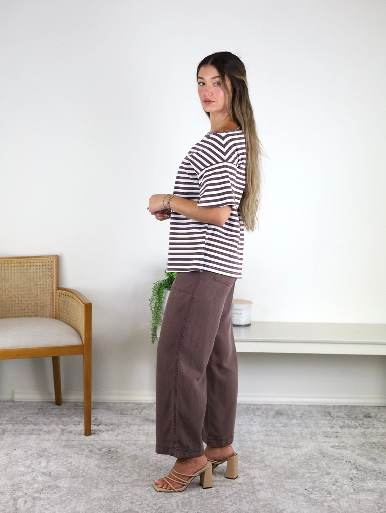 A woman stands sideways in a room, wearing the Hayden Asymmetrical Off-Shoulder Boxy Cotton Tee paired with brown pants and heeled sandals. In the background, there is a wooden chair and a small table with a plant.