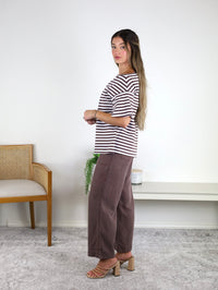 A woman stands sideways in a room, wearing the Hayden Asymmetrical Off-Shoulder Boxy Cotton Tee paired with brown pants and heeled sandals. In the background, there is a wooden chair and a small table with a plant.