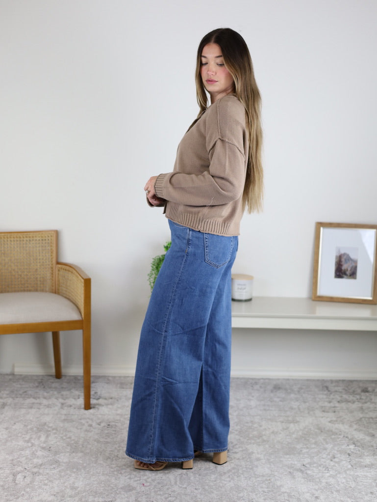 A person wearing a tan long-sleeved Jude Raw Edge Cardigan and wide-leg blue jeans.