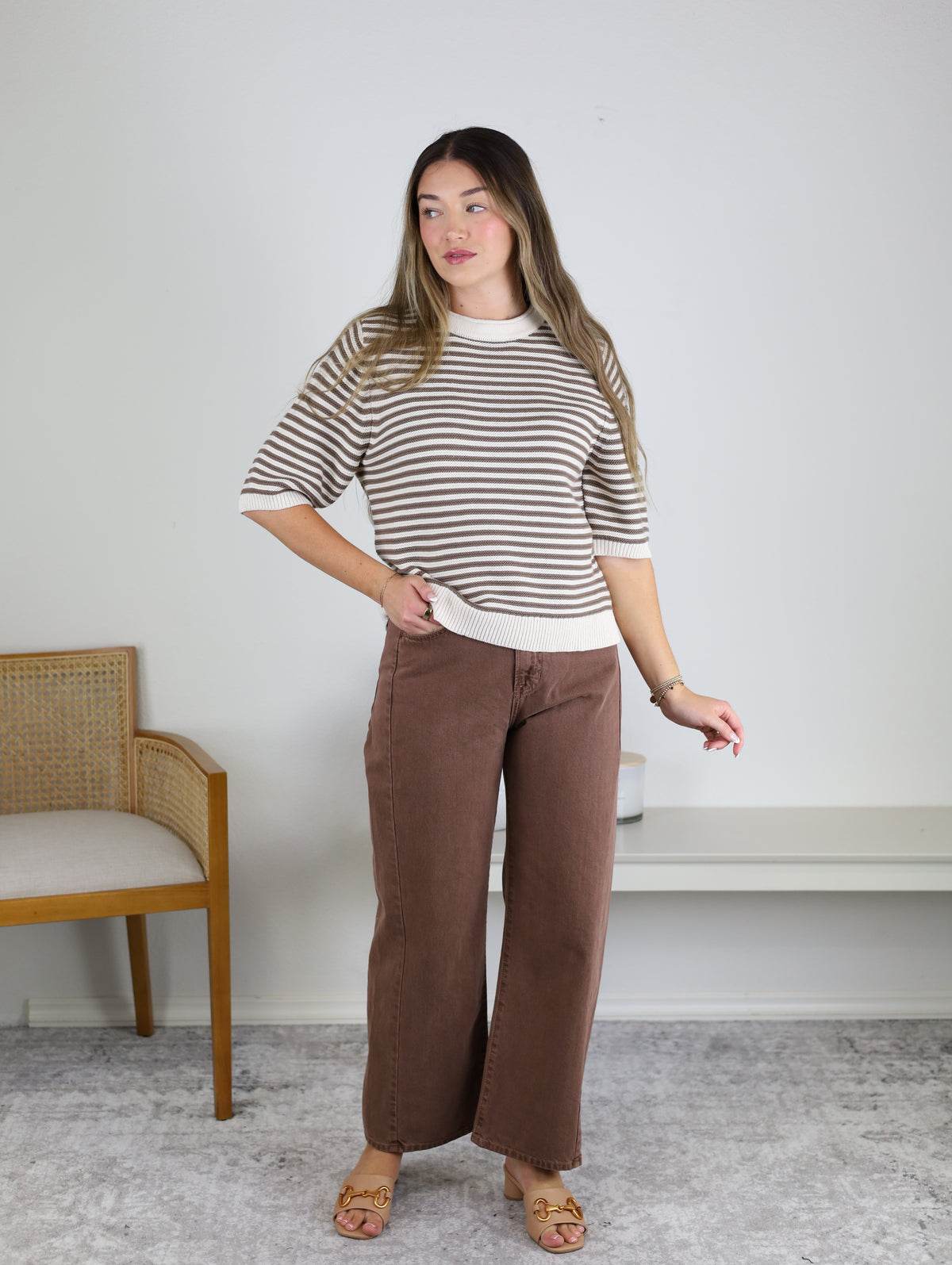 A woman with long hair wears the Scout Striped Sweater—beige and white striped cotton knit—paired with wide-legged brown pants. She stands indoors near a wooden chair and a white bench on a light gray carpet, looking to the side with one hand slightly raised.
