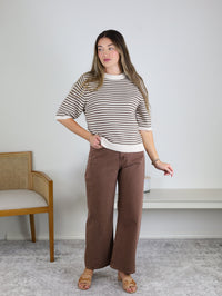 A woman with long hair wears the Scout Striped Sweater—beige and white striped cotton knit—paired with wide-legged brown pants. She stands indoors near a wooden chair and a white bench on a light gray carpet, looking to the side with one hand slightly raised.