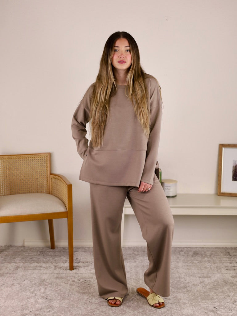 A woman wearing a taupe Arbor Lounge Set consisting of a matching long-sleeved top and wide-leg trousers.