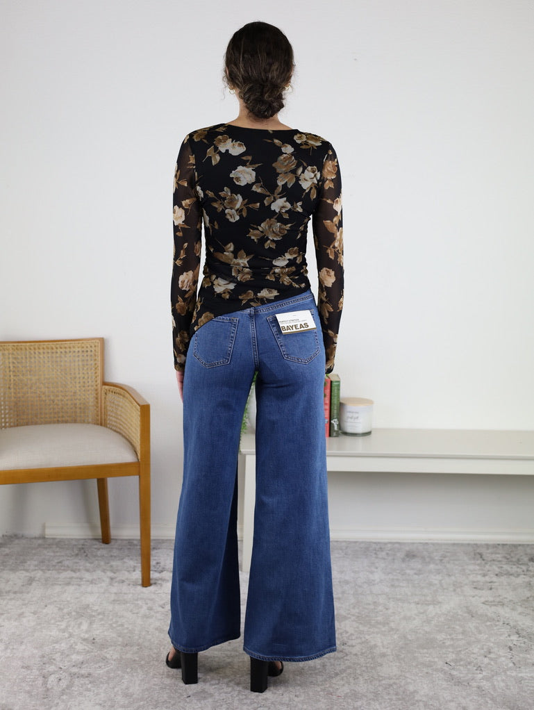 A person wearing wide-leg blue jeans and the Nightfall Rose Floral Mesh Asymmetrical Top, featuring long sheer mesh sleeves and a black floral design, stands facing away in a room with a chair, books, and decorative items on a white shelf.