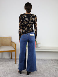 A person wearing wide-leg blue jeans and the Nightfall Rose Floral Mesh Asymmetrical Top, featuring long sheer mesh sleeves and a black floral design, stands facing away in a room with a chair, books, and decorative items on a white shelf.