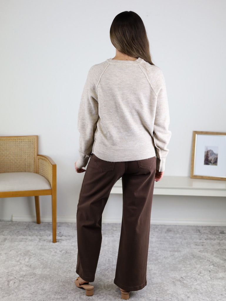 A woman with long brown hair, wearing the Theo Sweater and brown pants, stands indoors with her back to the camera. A wooden chair and a white table with a framed picture are beside her.