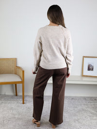 A woman with long brown hair, wearing the Theo Sweater and brown pants, stands indoors with her back to the camera. A wooden chair and a white table with a framed picture are beside her.