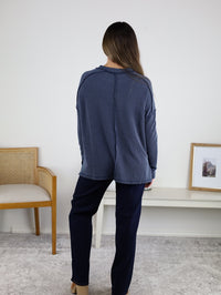 A woman with long hair, dressed in a loose blue Finley Sweater and navy pants, stands facing away in a minimal room with a chair, table, and small framed picture on the wall.