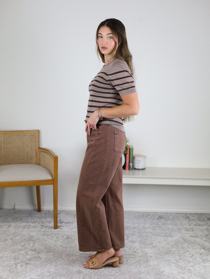 A woman in a brown striped shirt and the Raj High Rise Twist Seam Barrel Jean stands sideways in a minimal room with a chair, books, and decor on a shelf behind her.