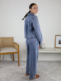 A woman with a braid, wearing the Adalyn Acid Wash Denim Boxy Shirt and jeans, stands sideways and smiles indoors near a chair and a white shelf adorned with a framed photo and candles.