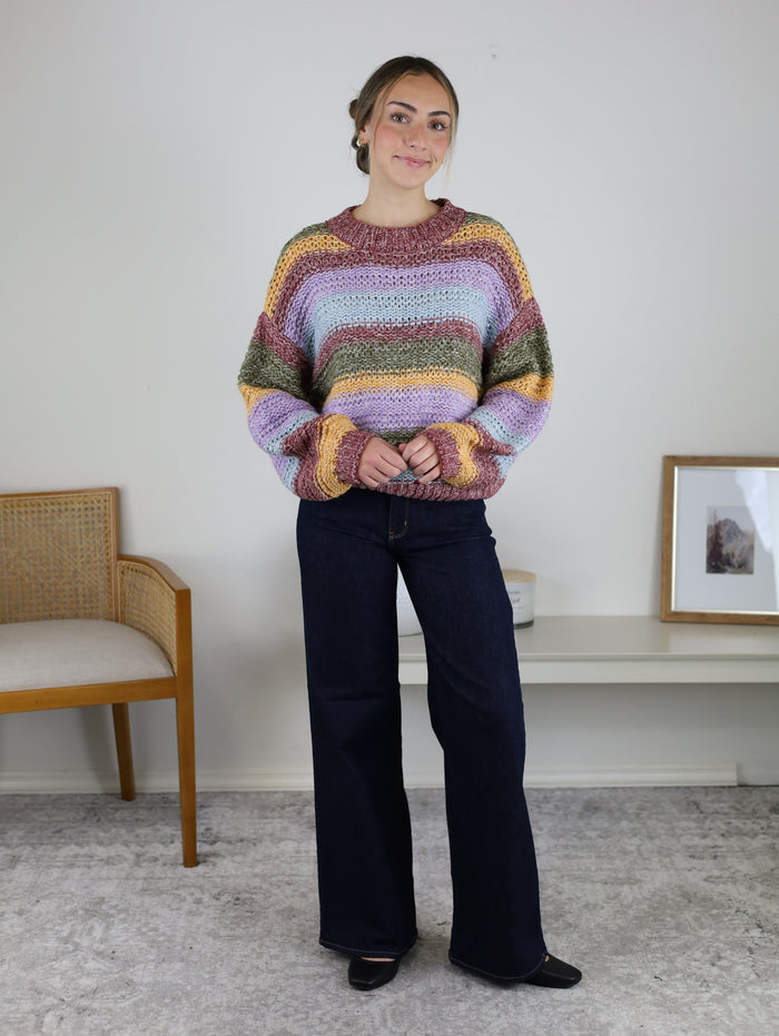 A person wears a Reign Striped Knit Sweater in shades of pink, lavender, green, and yellow, paired with dark wash wide-leg jeans.