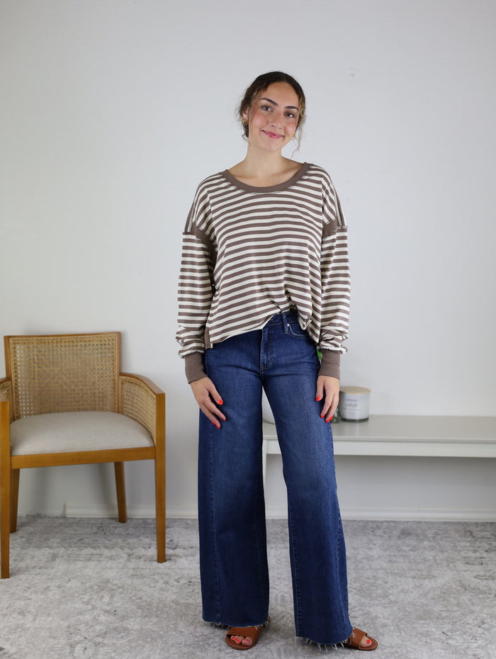 A woman stands indoors on a light carpet, smiling as she poses in the Kiana Stripe Open Back Long Sleeve top with brown and white stripes, wide-leg blue jeans, and sandals. She is next to a wooden chair and a white table against a plain wall.