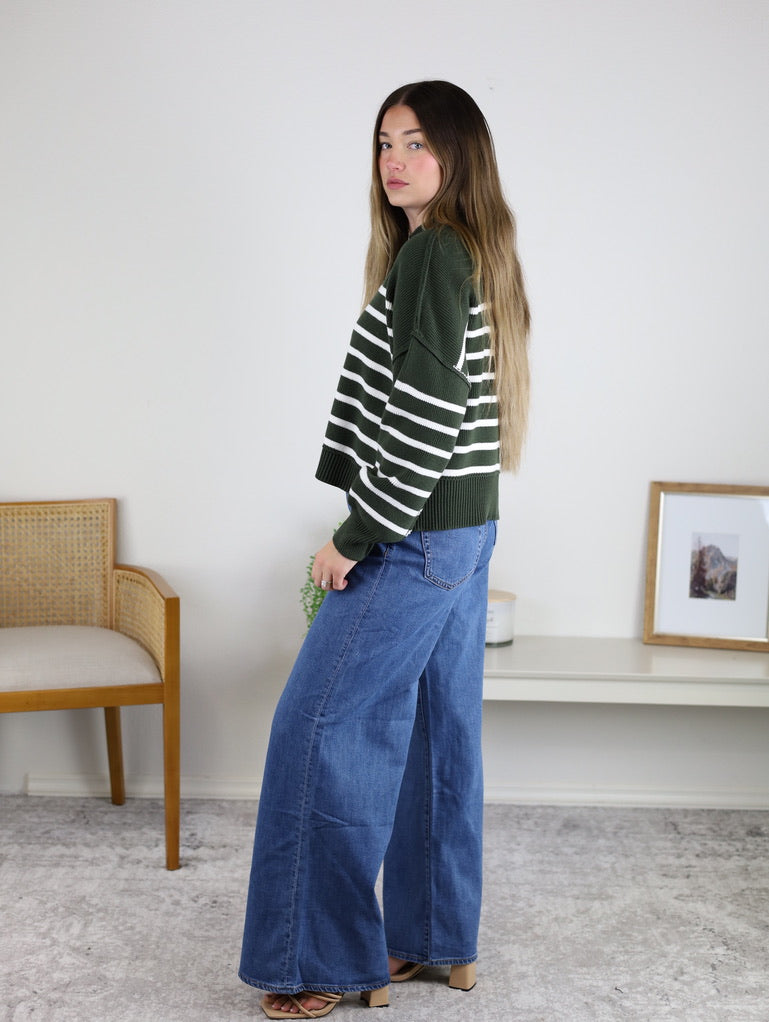 A woman wearing an oversized, dark green and white Noa Striped Sweater paired with wide-leg blue jeans.