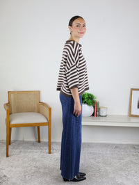 A woman stands sideways, wearing the Stephanie Striped Cardigan in chocolate brown over blue jeans. She smiles slightly with her hands relaxed by her sides, while a chair and decor can be seen in the background on a light-colored rug.