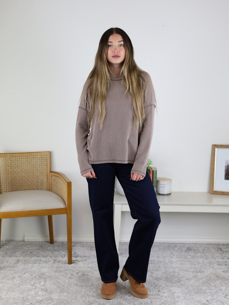 A woman with long hair wears a taupe ribbed knit Finley Sweater, dark pants, and brown shoes, standing in a room with a chair, white table, books, framed art, and a light rug.
